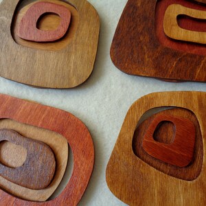 Op de afbeelding: Vier houten onderzetters met een concentrisch cirkelontwerp. De onderzetters zijn gemaakt van verschillende tinten bruin hout, met de binnenste cirkels geschilderd in tinten rood en bruin.
