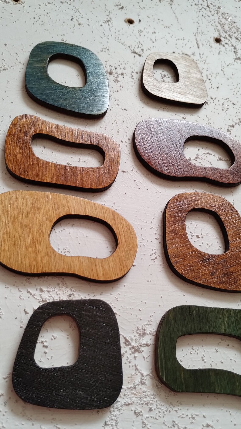 Op de afbeelding: Een set van acht houten wanddecoraties in verschillende vormen en maten. De stukken zijn gebeitst in tinten bruin, groen en zwart. Elk stuk heeft een uitsparing in het midden, waardoor een uniek ontwerp ontstaat.