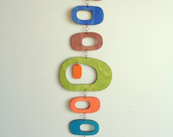 May include: A colorful wooden mobile with geometric shapes in shades of orange, blue, green, yellow, and brown. The shapes are suspended from a string and hang in a vertical line.