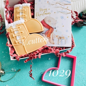 May include: Two gingerbread cookies shaped like winter boots in a white box with red and white tissue paper. A card with a polar bear and the text "Merry Christmas" is in the box. A pink cookie cutter shaped like a boot with the number 1029 is on a turquoise surface.