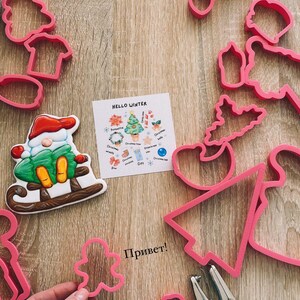 May include: A set of pink plastic cookie cutters in various shapes, including a Christmas tree, a snowman, a gingerbread man, and a star. The cutters are arranged on a wooden surface with a card that says "Hello Winter" and a list of Christmas-themed cookie cutter shapes. The word "Привет!" is written in black on the surface.