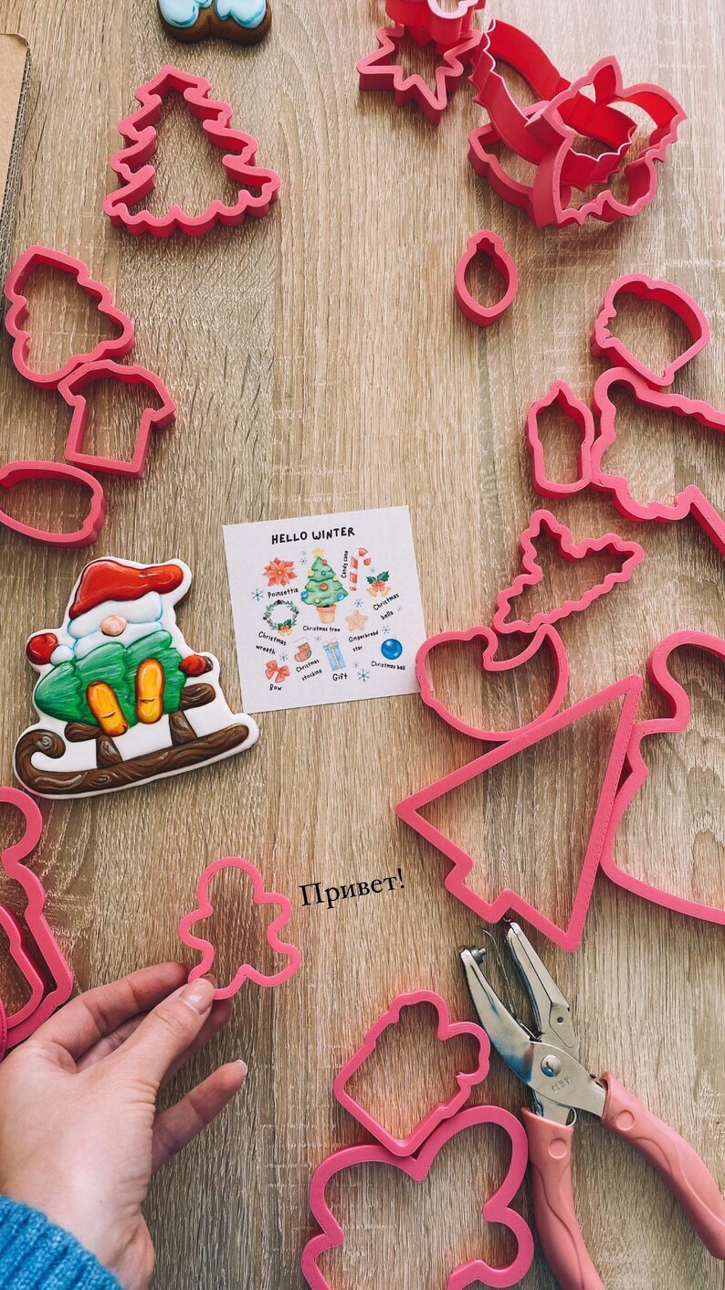 May include: A set of pink plastic cookie cutters in various shapes, including a Christmas tree, a snowman, a gingerbread man, and a star. The cutters are arranged on a wooden surface with a card that says "Hello Winter" and a list of Christmas-themed cookie cutter shapes. The word "Привет!" is written in black on the surface.