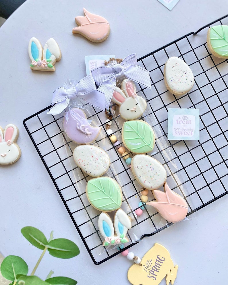 K&ouml;nnte beinhalten: Ein Set Oster-Zuckerkekse, verziert mit Pastellfarben und Fr&uuml;hlingsmotiven. Die Kekse sind in durchsichtigen Cellophanbeuteln verpackt, die mit wei&szlig;em Band gebunden sind. Die Beutel sind auf einem schwarzen Drahtgestell angeordnet. Ein Schild lautet "Ein kleiner Leckerbissen f&uuml;r jemanden, der s&uuml;&szlig; ist".