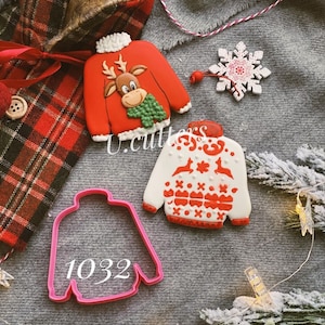 May include: Three Christmas cookie cutters in the shape of jumpers. One cutter is red with a reindeer design, one is white with a red and white pattern, and one is pink with the number 1032 printed on it.