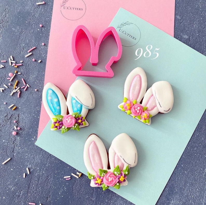 K&ouml;nnte beinhalten: Drei Hasen-f&ouml;rmige Zuckerkekse, verziert mit rosa und blauem Zuckerguss und bunten Blumen. Ein rosa Plastik-Ausstecher in Form von Hasenohren ist ebenfalls abgebildet. Der Ausstecher ist mit "U. Cutters" und der Nummer "985" beschriftet.