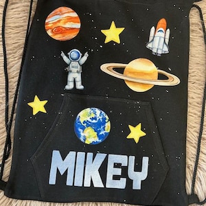 Może przedstawiać: Czarny plecak ze sznurkiem ze wzorem kosmicznym. Zawiera planety, gwiazdy, rakietę i astronautę. Imię "MIKEY" jest wydrukowane dużymi, jasnoniebieskimi literami na przedniej kieszeni. Torba ma czarne sznurki.