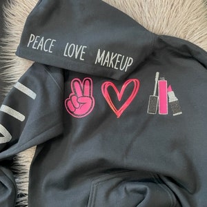 May include: Black hoodie with a silver glitter print that says "Peace Love Makeup" with a pink peace sign, heart, and lipstick graphics.