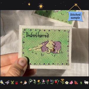 May include: A cross-stitched patch with the word "Unbothered" and an illustration of a character relaxing. The patch is green with a white border and a blue tag that says "Stitched sample".