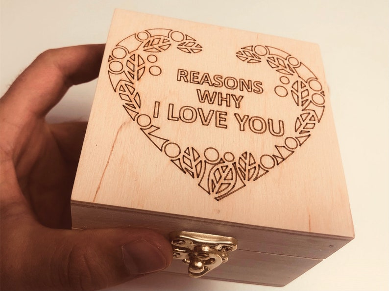 以下が含まれることがあります： 明るい色の木製ボックス。蓋には装飾的なハートのデザインと「REASONS WHY I LOVE YOU」の文字が刻まれています。ボックスには金色の留め金が付いています。