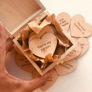 以下が含まれることがあります： 木製の箱と、ハート型の木製トークン。トークンには「You're my person」や「The trust between us」などのフレーズが刻まれています。箱は開いており、中身と箱を持っている手が見えます。