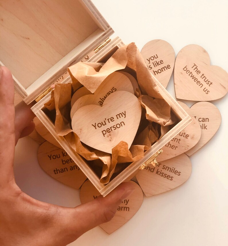 以下が含まれることがあります： 木製の箱と、ハート型の木製トークン。トークンには「You're my person」や「The trust between us」などのフレーズが刻まれています。箱は開いており、中身と箱を持っている手が見えます。