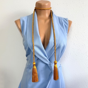 Könnte beinhalten: Ein goldfarbenes Abschluss-Ehrenband mit passendem Choker und zwei Quasten. Das Band ist über einem hellblauen ärmellosen Blazer auf einer Schaufensterpuppe drapiert. Die Quasten sind in einem dunkleren Goldton gehalten.