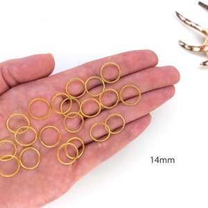 May include: A hand holding a pile of 14mm gold jump rings. The rings are all the same size and are arranged in a pile. The text "14mm" is visible in the lower right corner of the image.