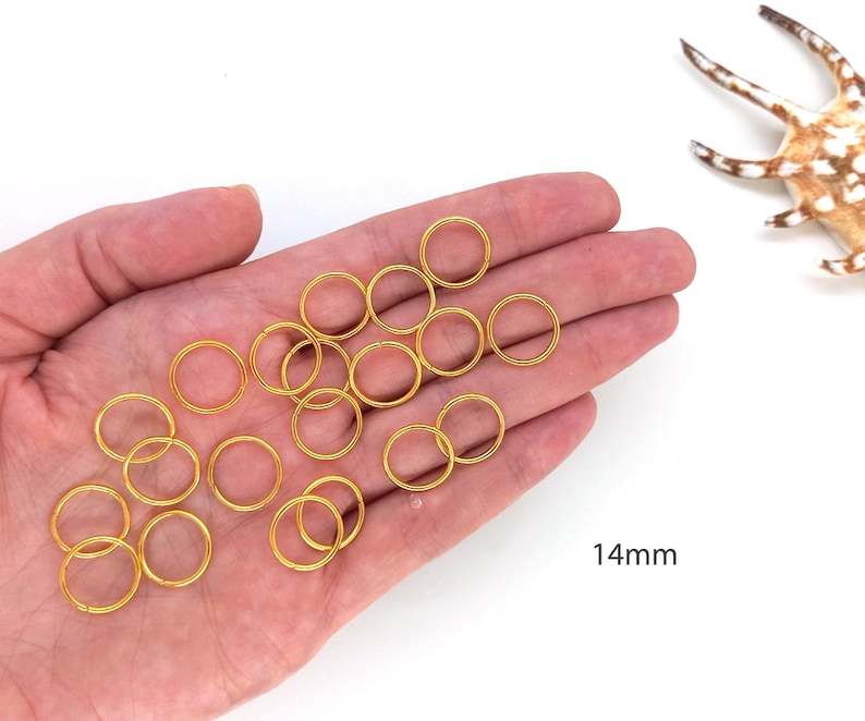 May include: A hand holding a pile of 14mm gold jump rings. The rings are all the same size and are arranged in a pile. The text "14mm" is visible in the lower right corner of the image.