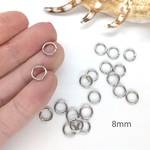May include: Silver jump rings, 8mm in diameter, scattered on a white surface. Some rings are held in a hand.