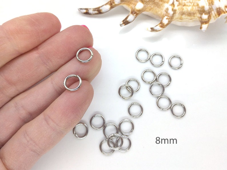 May include: Silver jump rings, 8mm in diameter, scattered on a white surface. Some rings are held in a hand.