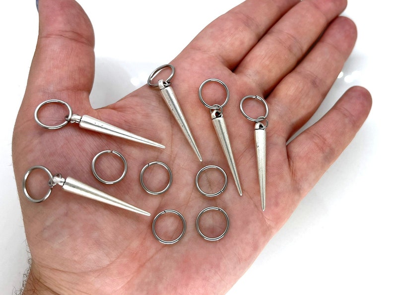 K&ouml;nnte beinhalten: Ein Set aus sechs silberfarbenen Nasenringen mit spitzem Design. Die Ringe werden auf einer Hand gezeigt, wobei f&uuml;nf der Ringe ein Reifen-Design und ein Ring ein Schl&uuml;sselring-Design haben.