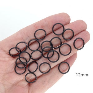 May include: A hand holding a pile of black metal jump rings, each measuring 12mm in diameter.