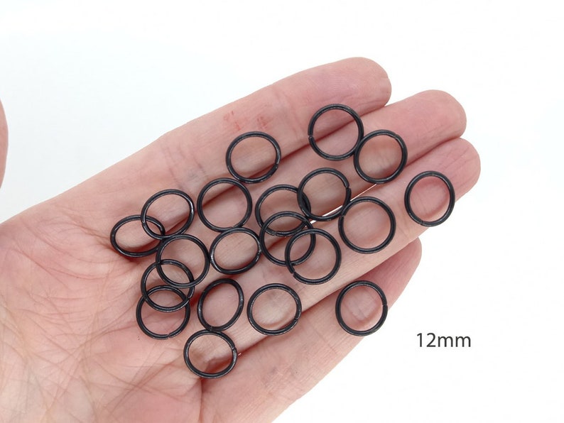 May include: A hand holding a pile of black metal jump rings, each measuring 12mm in diameter.