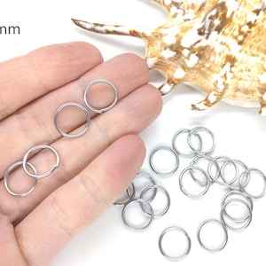May include: A close-up of a hand holding a pile of silver jump rings, each measuring 12mm in diameter. The jump rings are arranged in a loose pile, with some overlapping. The background is a white surface.