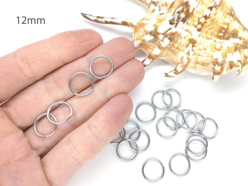 May include: A close-up of a hand holding a pile of silver jump rings, each measuring 12mm in diameter. The jump rings are arranged in a loose pile, with some overlapping. The background is a white surface.