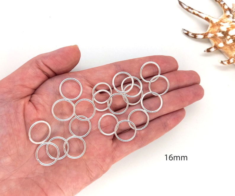 May include: A hand holding a pile of silver jump rings, each measuring 16 mm in diameter.