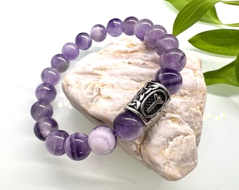 May include: A beaded bracelet featuring round, polished amethyst stones. The bracelet has a silver-colored cylindrical accent bead with an engraved design. The purple stones are approximately 0.8 cm in diameter. The bracelet is displayed on a light-colored stone.