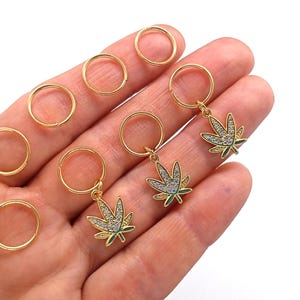 May include: Gold-colored hoop earrings and charms. The charms are shaped like cannabis leaves, with clear crystal accents and green enamel detailing. Several plain gold hoops are also visible.