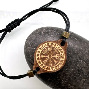 Può includere: Un braccialetto in corda nera con un ciondolo in legno che presenta una bussola e rune. Il ciondolo è marrone con accenti dorati.
