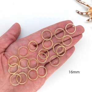 May include: A close-up of a hand holding 16mm gold jump rings. The rings are arranged in a loose pile, with some overlapping. The background is white.