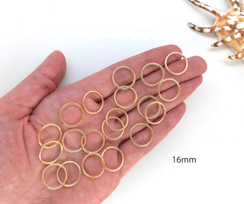 May include: A close-up of a hand holding 16mm gold jump rings. The rings are arranged in a loose pile, with some overlapping. The background is white.