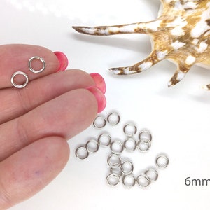 May include: Silver jump rings, 6mm in diameter, scattered on a white surface. There are two jump rings in a hand, showing the size.