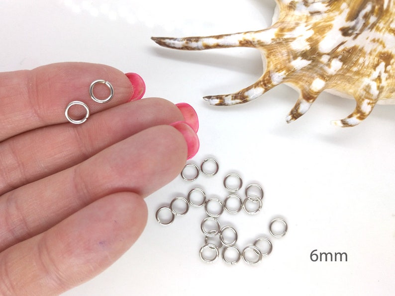 May include: Silver jump rings, 6mm in diameter, scattered on a white surface. There are two jump rings in a hand, showing the size.