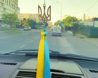 Może przedstawiać: Żółto-niebieski frędzl zwisa z lusterka wstecznego w samochodzie. Frędzl jest przymocowany do złotego metalowego symbolu ukraińskiego trójzębu.