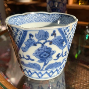 May include: A small, white ceramic cup with a blue and white floral design. The cup has a scalloped rim and a geometric pattern around the base.