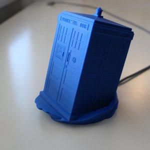 May include: A blue 3D printed model of a police box with the text "Police Box" on the side. The model is sitting on a blue base.