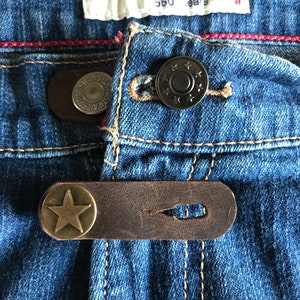 May include: Close-up of a pair of blue denim jeans with a brown leather belt loop and a star-shaped metal button. The jeans have a red stitching detail around the waistband.