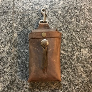 May include: A brown leather phone case with a silver clip and a leather strap. The case has a worn look and a distressed finish.