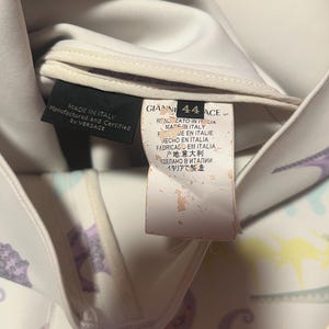 May include: Close-up of a white garment with a colorful pattern and multiple tags. One tag reads "MADE IN ITALY" and "Manufactured and Certified by VERSACE." Another tag displays "GIANNI 44 ACE" and text in multiple languages, including Italian and Japanese.