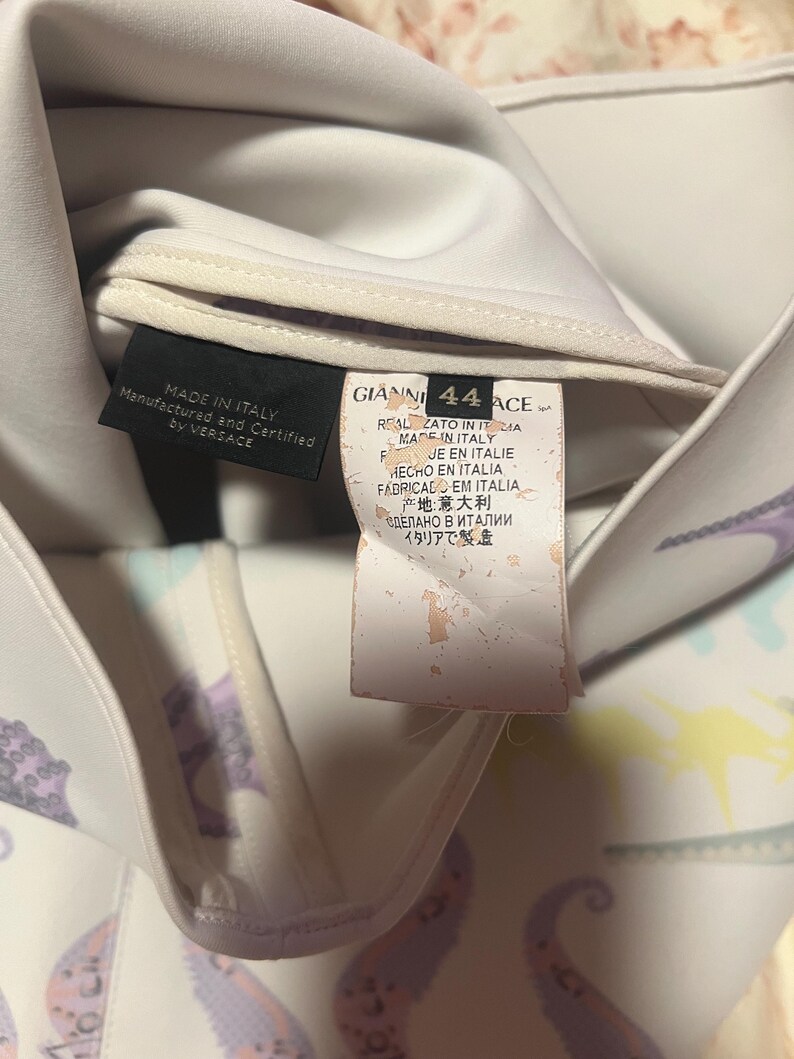 May include: Close-up of a white garment with a colorful pattern and multiple tags. One tag reads "MADE IN ITALY" and "Manufactured and Certified by VERSACE." Another tag displays "GIANNI 44 ACE" and text in multiple languages, including Italian and Japanese.