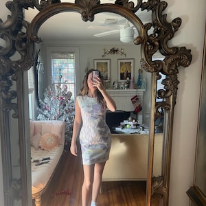 May include: A sleeveless, bodycon dress with a pastel floral pattern. The dress is white with a design in shades of purple, blue, and yellow. The person in the image is wearing white socks. The dress is reflected in a large, ornate, gold-framed mirror.