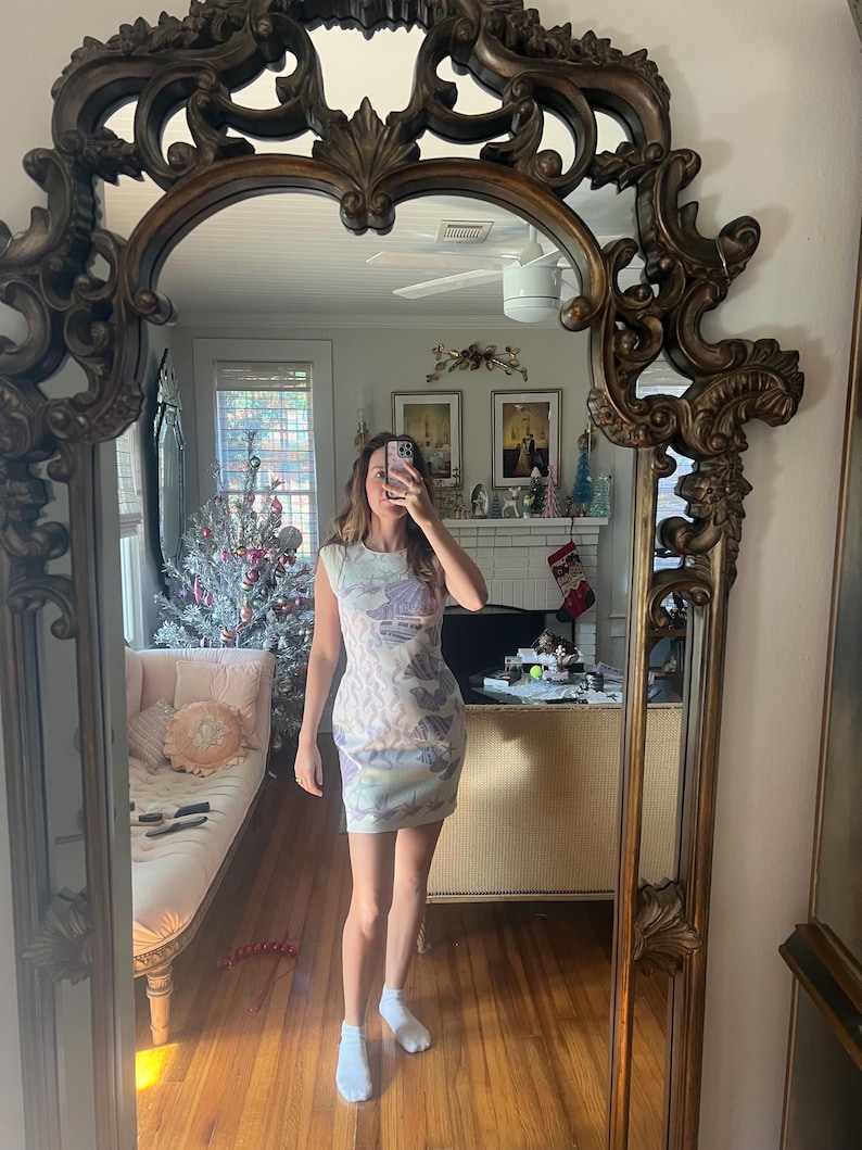 May include: A sleeveless, bodycon dress with a pastel floral pattern. The dress is white with a design in shades of purple, blue, and yellow. The person in the image is wearing white socks. The dress is reflected in a large, ornate, gold-framed mirror.