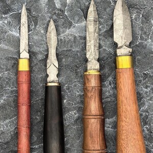 May include: Four handcrafted knives with unique blade designs and wooden handles. The handles vary in color and shape, including a cylindrical red handle, a black handle with a green band, and two brown wooden handles. The blades have a textured pattern.