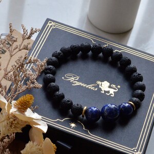 May include: A black lava rock bracelet with a gold accent and three blue lapis lazuli beads. The bracelet is in a black box with gold lettering that says "Regulus".