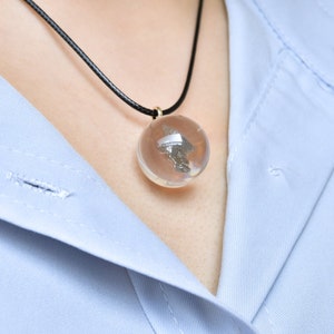 May include: A silver pendant necklace with a clear glass orb containing a small piece of metal. The necklace is on a black cord and is worn by a person with a light blue shirt.
