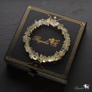 Puede incluir: Un brazalete de chips de cuarzo transparente en una caja negra con detalles dorados. La caja tiene la palabra "Regulus" impresa en letras doradas.