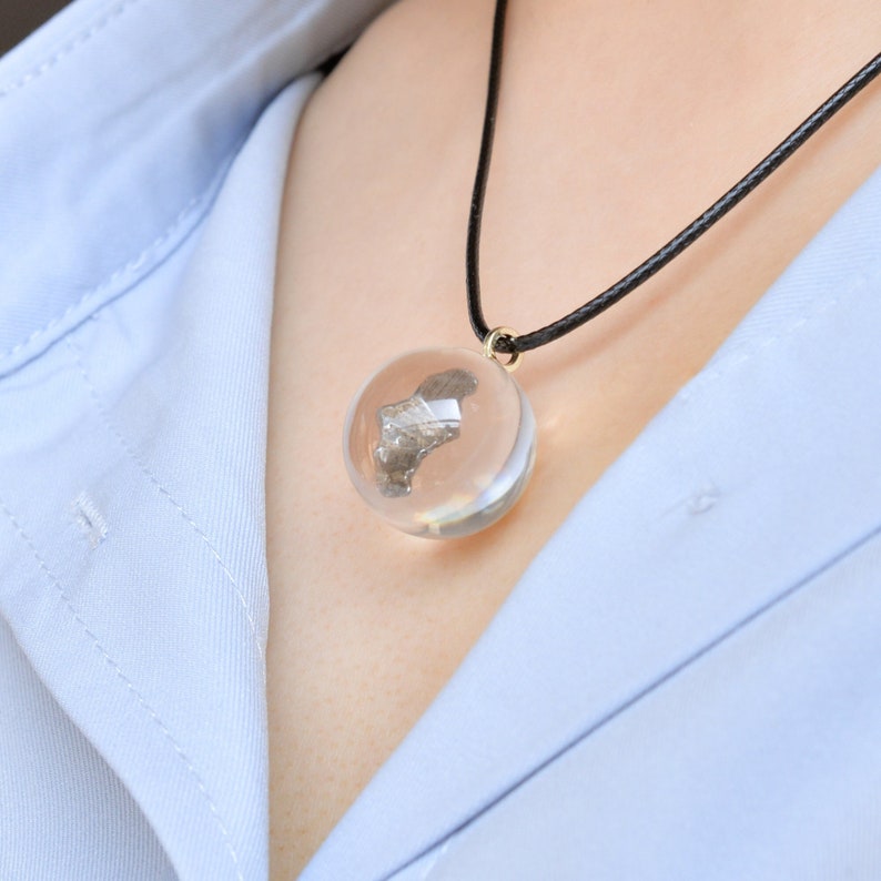 May include: A silver-colored metal object encased in a clear glass sphere pendant on a black cord necklace.