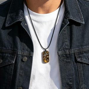 Puede incluir: Collar con colgante rectangular y cord&oacute;n negro. El colgante presenta un dise&ntilde;o marr&oacute;n y amarillo en un marco de metal plateado. La persona lleva una camiseta blanca y una chaqueta vaquera azul oscuro.