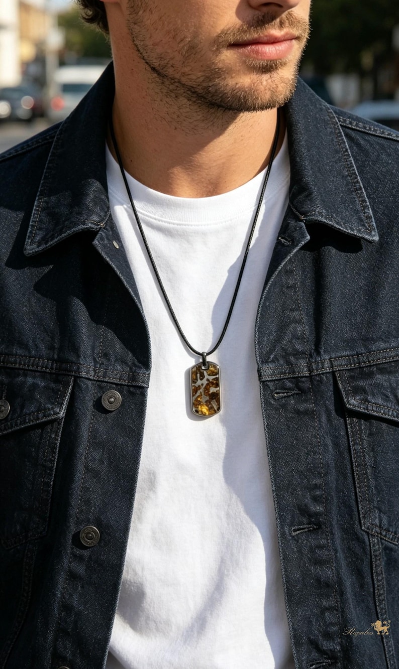 Puede incluir: Collar con colgante rectangular y cord&oacute;n negro. El colgante presenta un dise&ntilde;o marr&oacute;n y amarillo en un marco de metal plateado. La persona lleva una camiseta blanca y una chaqueta vaquera azul oscuro.