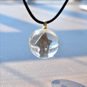 May include: A silver-colored star-shaped pendant encased in a clear glass sphere, hanging from a black cord necklace.